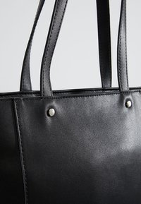 Czarna torba typu tote wykonana ze skóry, wyposażona w dwa uchwyty, teksturowaną powierzchnię, srebrne elementy dekoracyjne oraz widoczne przeszycia.