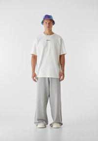 Biela voľná tričko s potlačou "STRATENÁ MLÁDEŽ", šedé teplákové nohavice a viacfarebný klobúk s okrajom. Má široké nohy a slip-on topánky.