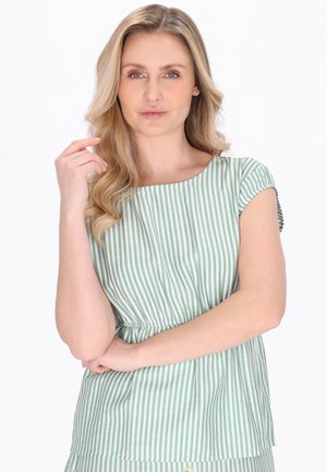 Kvinde med langt blondt hår i en grøn- og hvidstribet kortærmet top, poserer med den ene arm over kors og den anden hånd nær ansigtet.