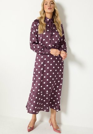 Femme en robe midi violette à manches longues avec des pois blancs, debout, portant des talons pointus mauve avec des boucles décoratives.