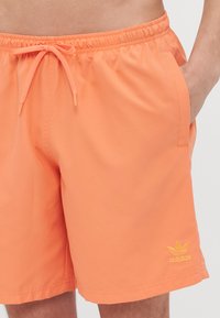 Man wearing orange Adidas swim shorts with a drawstring waist and side pockets, hand partially in right pocket.