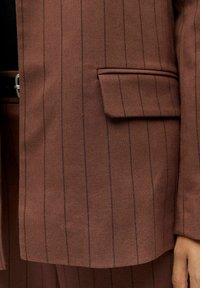 Gros plan sur la poche d'un blazer marron à rayures avec rabat et un pantalon assorti, vue partielle d'une main et d'une boucle de ceinture sur le côté.