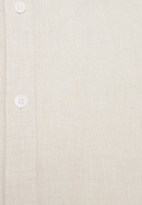 Camicia button-up beige con trama a rete, caratterizzata da bottoni bianchi e dettagli di cucitura visibili. Tessuto liscio con un aspetto morbido.