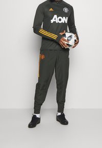 Grå sportsoverall med orange accenter, långärmad och med hög krage, i kombination med svarta skor. Personen håller i en mönstrad fotboll.