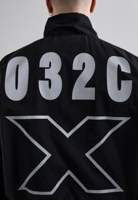 Chaqueta negra con gran texto blanco "032C" encima de un diseño de "X" en negrita en la parte trasera. El material parece suave con un acabado mate.