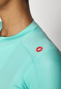 Turkizna sportska majica s finim rebrastim uzorkom, visokim izrezom i crvenim kružnim logom na ramenu. Prikladan dizajn za aktivno nošenje.