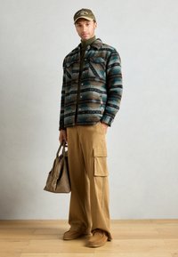Jakna od vune s uzorkom u plavoj, smeđoj i crnoj boji; kašmirster turtleneck u boji khaki; bež cargo hlače; smeđa torba; bež obuća; kapa s tekstualnim detaljem.