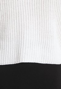 Pull en maille côtelée blanche avec un ourlet noir, présentant une finition texturée et un design court. Le tissu est doux avec des détails de coutures visibles.