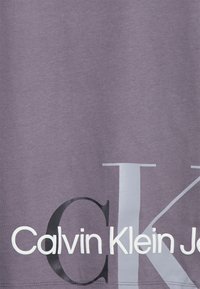 Camiseta de algodón púrpura con gran texto en blanco y negro: "Calvin Klein Jeans" con una letra "K" prominente. Textura suave.