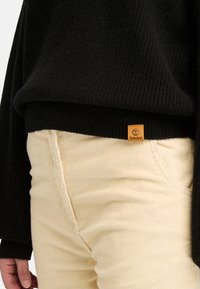 Svart stickad tröja med ribbade ärmmuddar, kombinerad med krämfärgade manchesterbyxor. Litet brunt Timberland-logomärke synligt.