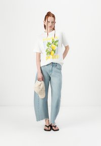 T-shirt bianco in cotone con grafica "SOUR & SWEET", abbinato a jeans a gamba larga azzurri, sandali neri e una borsa all'uncinetto.