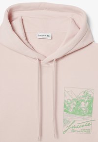 Sudadera rosa con cordones, que presenta un gráfico verde de montañas y árboles etiquetado "Lacoste Tennis Heritage". Textura acanalada en el interior.