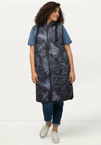 Gep puffter ärmelloser marineblauer Weste mit strukturiertem Muster, Frontreißverschluss und Kordelzugkapuze. Tragen über einem Jeanshemd und gestylt mit Jeans.