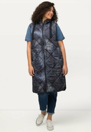 Gilet sans manches matelassé bleu marine avec un motif texturé, fermeture éclair à l'avant et capuche avec cordon de serrage. Porté par-dessus une chemise en jean et associé à un jean.
