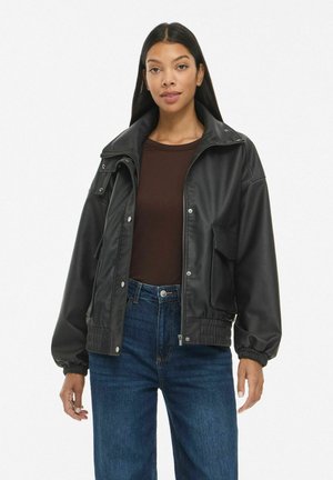 Femme aux longs cheveux foncés portant une veste en cuir noire, une chemise marron foncé et un jean bleu, debout devant un fond blanc uni.