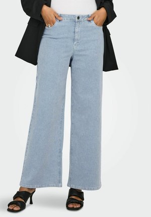 Személy világoskék, széles szárú farmernadrágot visel finom, függőleges csíkokkal, fekete magassarkú szandált, fekete blézert és fehér felsőt.