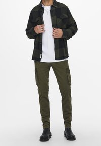 Camisa de franela a cuadros verde oscuro y negro sobre una camiseta blanca, combinada con pantalones cargo oliva y botines negros.