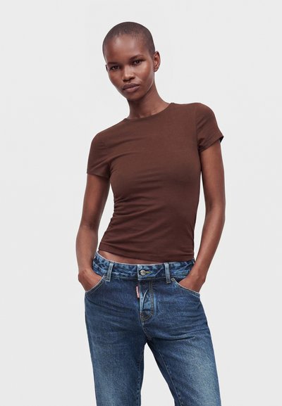 Femme aux cheveux courts portant un t-shirt marron ajusté à manches courtes et un jean bleu, les mains dans les poches, sur un fond uni.