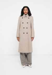 Cappotto beige in misto lana, doppiopetto, con grandi bottoni neri, colletto alto, vestibilità aderente e tasche laterali. Indossato sopra pantaloni a gamba larga a scacchi.