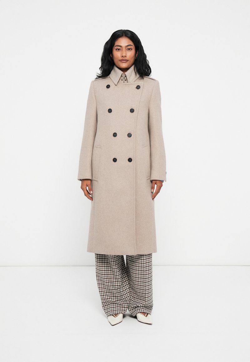 Cappotto beige in misto lana, doppiopetto, con grandi bottoni neri, colletto alto, vestibilità aderente e tasche laterali. Indossato sopra pantaloni a gamba larga a scacchi.