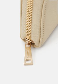 Borsa in tessuto beige con zip dorata, caratterizzata da una superficie texturizzata e una forma rettangolare. Accentuata con un punto esclamativo.
