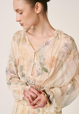 Mujer con el pelo recogido en un moño, vistiendo una blusa floral transparente con puños con volantes, sosteniendo pulseras doradas en sus manos contra un fondo liso.