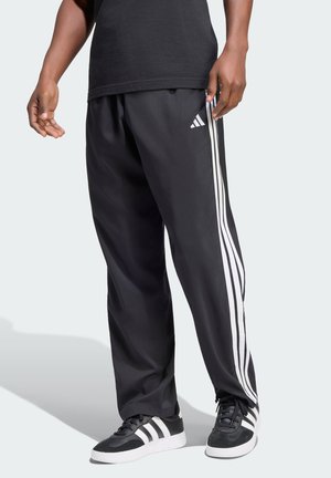 Osoba nosząca czarne spodnie dresowe Adidas z białymi paskami i pasujące czarno-białe sneakersy, stojąca na jasnym tle.