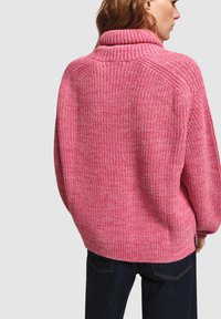 Roze gebreide trui met een hoge kraag, gestructureerde stof, verlaagde schouders en geribbelde zoom. Draagt over donkere spijkerbroeken, gezien van achteren.