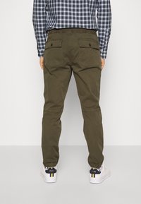 Pantalones caqui tapered con puños elásticos y bolsillos traseros, combinados con una camisa a cuadros en azul y blanco. El material parece suave y duradero.