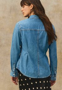 Lichte blauwe denimshirt met een klassieke kraag, lange mouwen met knoopmanchetten en een ronde zoom. De achterkant heeft een subtiele plooidetail.