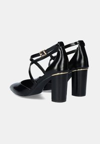 Sandalias de tacón alto de charol negro con punta afilada, diseño de tiras cruzadas y acento dorado en el tacón. Textura suave y construcción robusta.