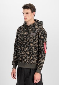 Giovane uomo con una felpa con cappuccio a stampa leopardata verde e nera e un'etichetta rossa "Rimuovere prima del volo" sul taschino della manica sinistra, in piedi su uno sfondo bianco.