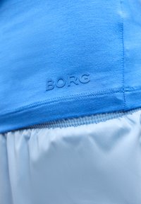 Μπλε υφασμάτινο τοπ με ραμμένο τελείωμα πάνω από ανοιχτό γκρι ρυτιδωμένο ύφασμα με ανάγλυφο κείμενο «BORG» κοντά στη ραφή.