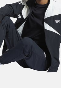 Person som bär en svart och vit Reebok träningsoverall sitter med ett knä upphöjt, vilket visar detaljerna på jackan och byxorna.