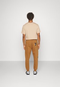 Man med lockigt hår står vänd ifrån, iklädd en beige t-shirt, bruna joggarbyxor med en lapp på bakfickan, och vita svarta Nike Air sneakers.