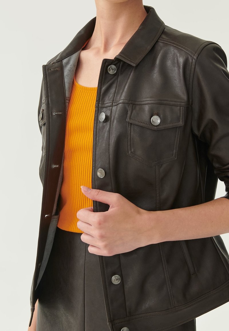 Giacca in pelle marrone scuro con colletto, bottoni a scatto e due tasche sul petto, indossata sopra un top arancione a coste. Texture liscia e design aderente.