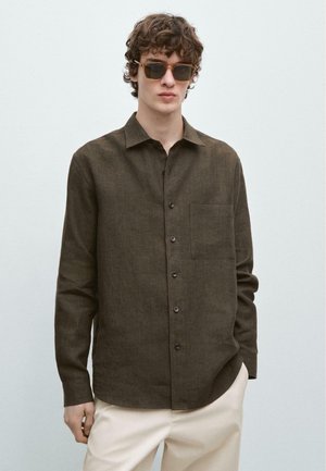 Junger Mann mit lockigem Haar, der ein braunes Hemd zum Knöpfen, beige Hosen und dunkle Sonnenbrillen trägt und vor einem schlichten hellen Hintergrund steht.