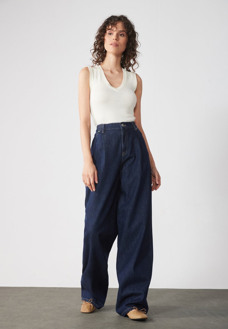 Krämfärgad ärmlös topp med djup V-ringning, matchad med oversized mörkblå denimbyxor. Byxorna har en strukturerad midja och vida ben.