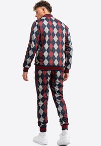 Rood, marineblauw en grijs ruitmotief trainingspak met geribde manchetten en kraag. Model wordt van achteren getoond, met witte sneakers.