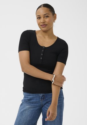 Kobieta z włosami związanymi do tyłu, ubrana w czarny top z krótkim rękawem i niebieskie dżinsy, trzymająca lewą rękę z srebrną bransoletką na nadgarstku.