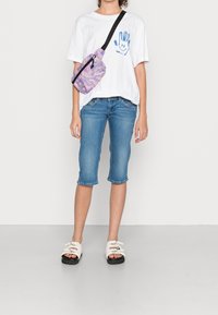 Persona che indossa una maglietta bianca con un sorriso a mano blu, pantaloni capri di denim blu, sandali bianchi e una borsa a tracolla viola tie-dye.