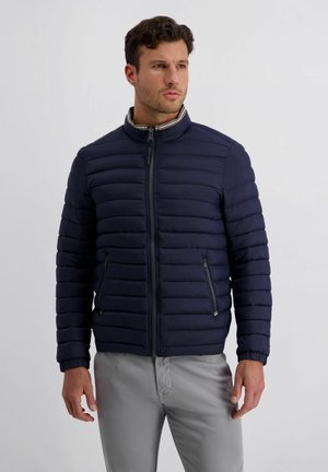 Man in een donkerblauwe doorgestikte jas met rits en opstaande kraag, en lichtgrijze broek, staande tegen een effen lichte achtergrond.