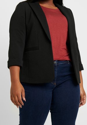 Femme portant un blazer noir texturé aux manches retroussées, un t-shirt rouge à col rond et un jean bleu foncé, devant un fond uni.