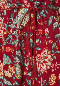 Robe fleurie rouge avec une taille nouée ornée de détails tressés et de pompons. Le tissu présente des motifs vibrants de fleurs et de feuilles en bleu et jaune.