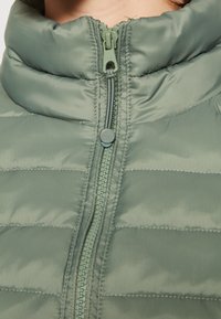 Jasnozielona kurtka puchowa z wysokim kołnierzem, z przodu zapinana na zamek błyskawiczny i poziomym przeszyciem. Materiał ma gładką, błyszczącą teksturę.