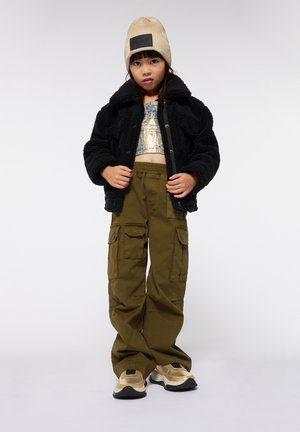Zwarte fleecejack, metallic crop top, olijfgroene cargo pants met zakken, en beige gebreide beanie. Gouden en zwarte accent sneakers.