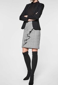 Svart kavaj, svart blus, grå rutmönstrad kjol med volanger och knähöga svarta strumpor. Kjolen har en kroppsnära passform och strukturerat mönster.