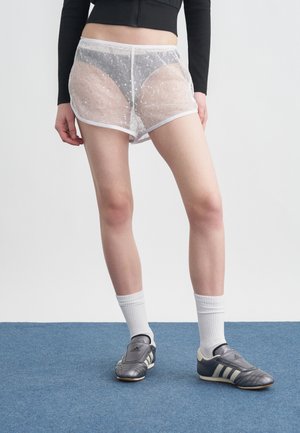 Persona che indossa pantaloncini bianchi a rete sopra biancheria intima nera, top nero a maniche lunghe, calzini bianchi e scarpe Adidas nere, in piedi su un tappeto blu.