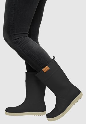 Person wearing black rubber rain boots with beige soles and black fitted jeans against a plain light background.