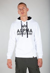 Sudadera blanca con cordones negros y logo "ALPHA INDUSTRIES". Cuenta con un bolsillo tipo canguro en la parte frontal y puños acanalados. Textura suave y lisa.